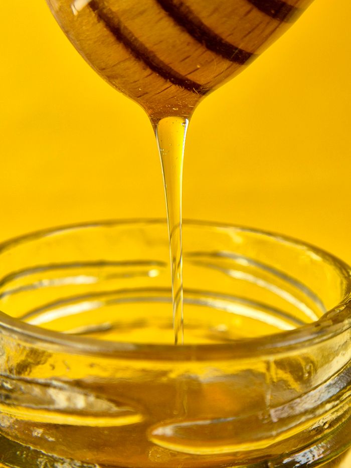 services-04 Close-up of golden honey flowing from dipper into a glass jar against a yellow background.