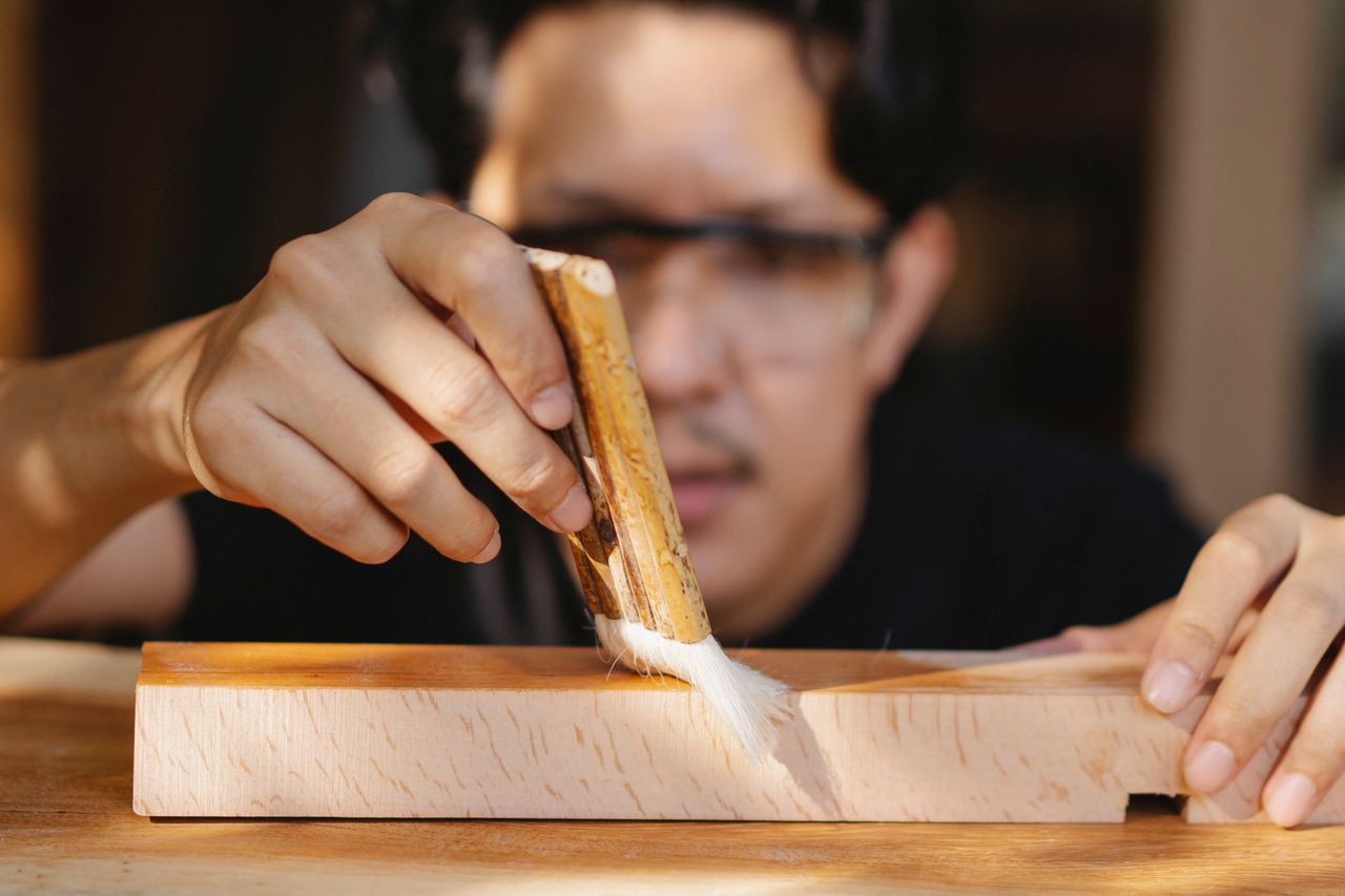 Young ethnic craftsman in casual clothes varnishing piece of wood with brush while working in workshop